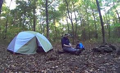 A picture of a dad with thier son sitting outside a tent in the forest.